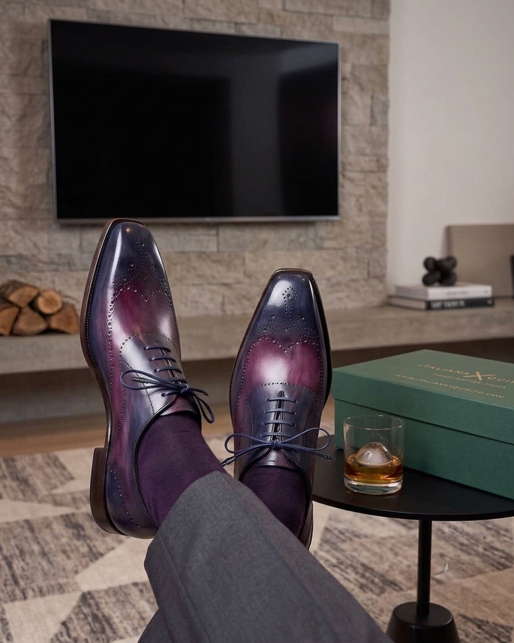 A pair of men's brogue dress shoes with a glossy purple and blue gradient finish, styled with dark purple socks and gray trousers. The shoes are shown with legs crossed in a modern living room setting next to a green box and a glass of whiskey.