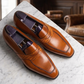 A pair of men's brown leather penny loafers with decorative brogue stitching and dark purple insoles, displayed on a white marble surface with a blurred office background.