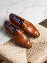 A pair of men's brown leather penny loafers with decorative brogue stitching and dark purple shoe trees inside, displayed on a woven rattan chair over a white marble surface.