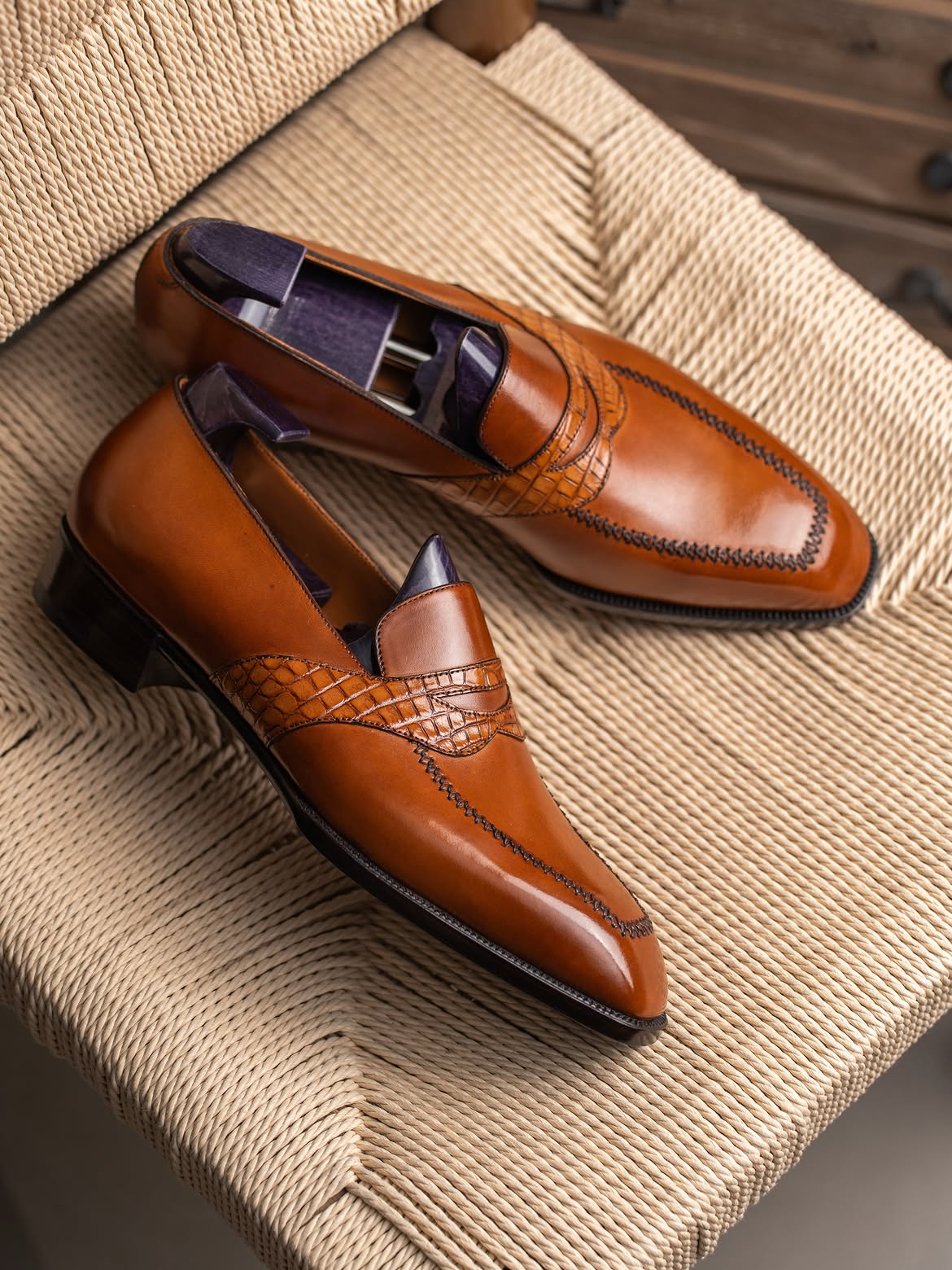 A pair of polished brown leather penny loafers with decorative stitching and embossed detailing, displayed on a woven beige chair.