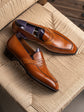 A pair of polished brown leather penny loafers with decorative stitching and embossed detailing, displayed on a woven beige chair.