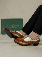 A person wearing brown and white wingtip oxford shoes with striped socks, with the green Italian X Quisito shoebox in the background.