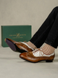 A person wearing brown and white wingtip oxford shoes with striped socks, with the green Italian X Quisito shoebox in the background.