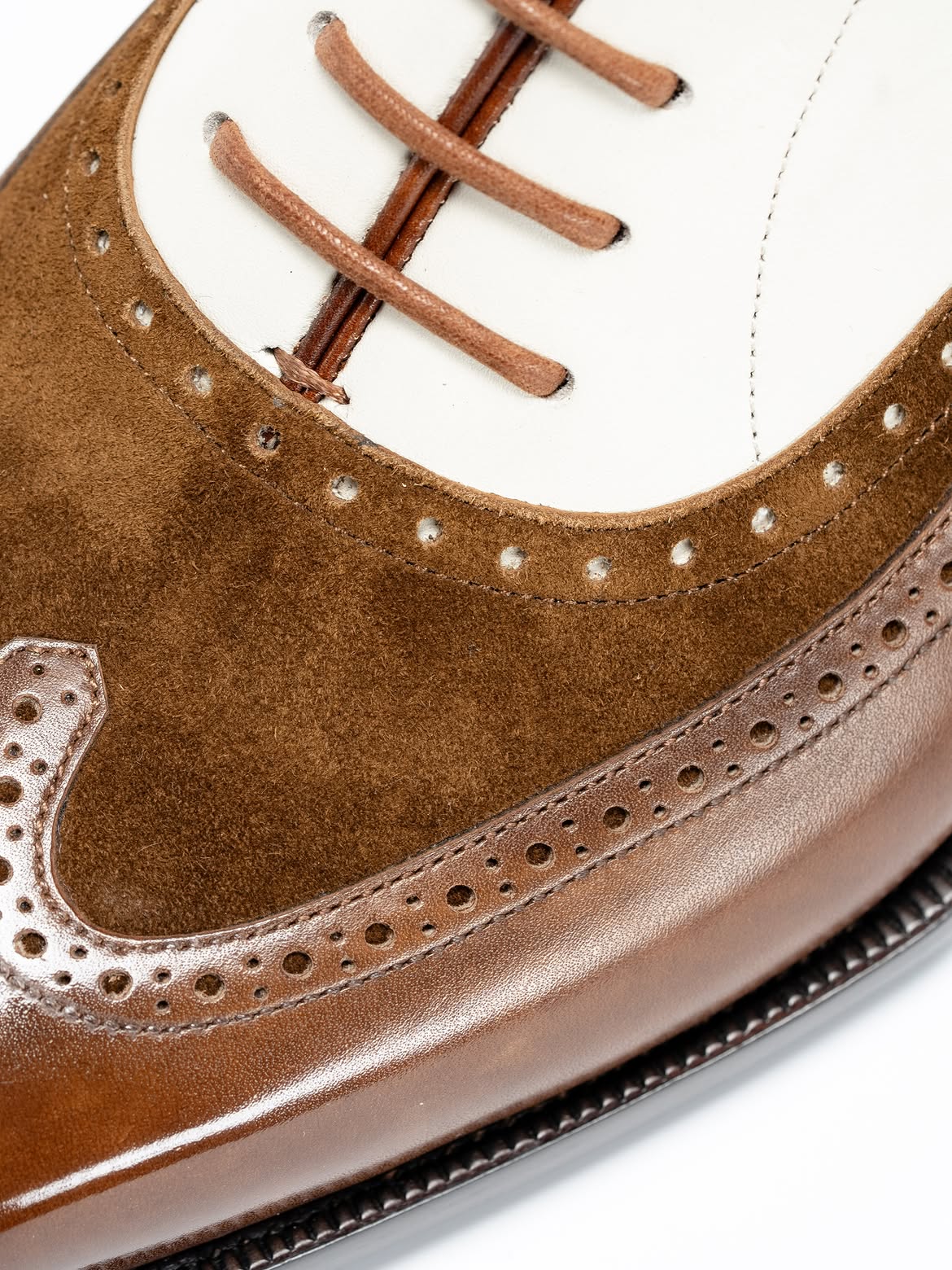 Close-up of a brown and white wingtip oxford shoe showing the suede vamp, leather toe cap, brogue perforations, and laces on a white background.