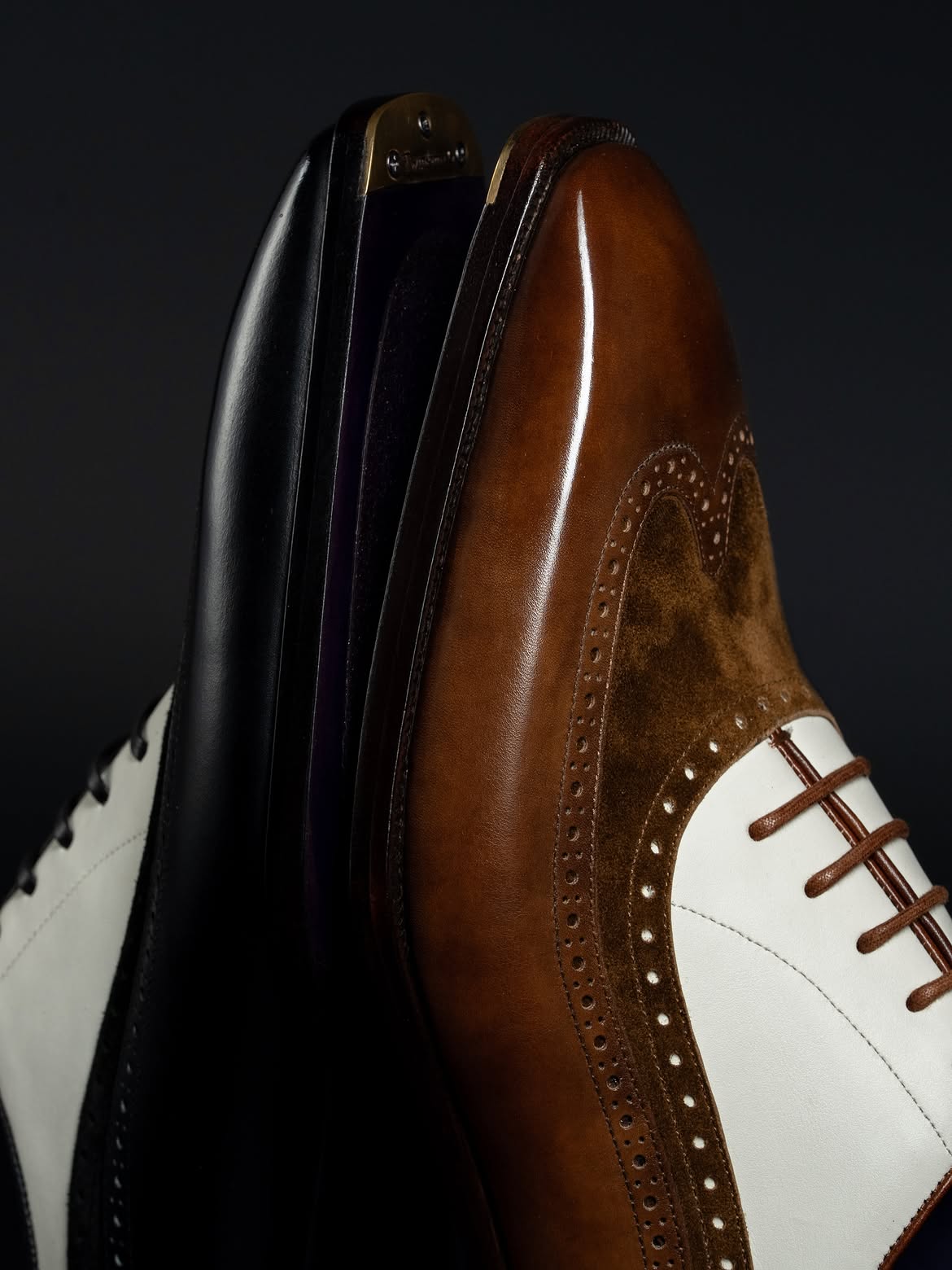 A close-up studio shot of a brown leather wingtip shoe with white side panels and dark brogue detailing, standing next to a black leather shoe against a dark background.