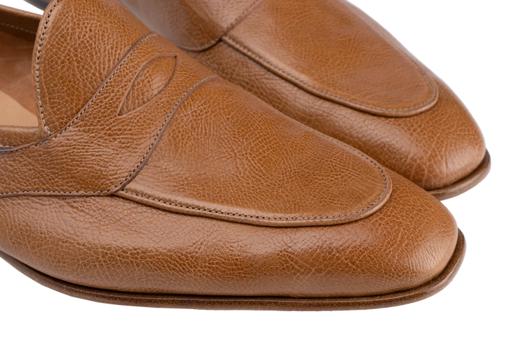 Close-up of a pair of tan leather penny loafers with a pebbled texture, showing the toe and side panels against a white background.