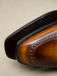 Close-up of a brown leather wingtip shoe showing the toe cap, brogue detailing, and dark sole edge against a neutral beige background.