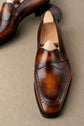 A pair of polished brown leather penny loafers with brogue detailing, shown on wooden shoe trees against a neutral beige background.