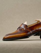 A close-up side view of a brown leather penny loafer with brogue detailing, shown on a light beige textured background.