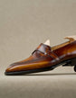 A close-up side view of a brown leather penny loafer with brogue detailing, shown on a light beige textured background.