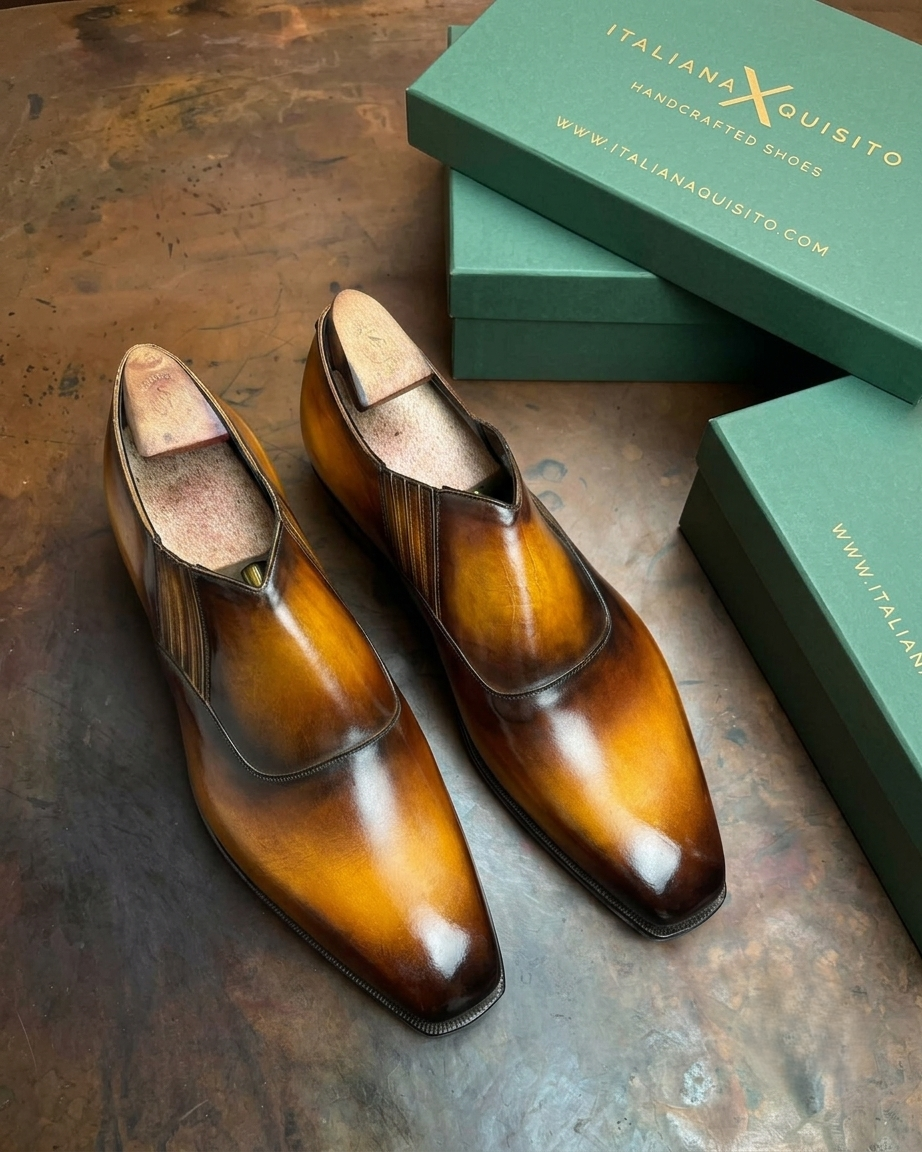 A pair of glossy brown leather dress shoes with a pointed toe and elastic side detailing, displayed next to green Italian Aquisito shoe boxes on a rustic dark metal surface.