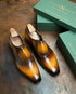 A pair of glossy brown leather dress shoes with a pointed toe and elastic side detailing, displayed next to green Italian Aquisito shoe boxes on a rustic dark metal surface.
