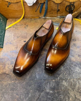 A pair of men's dress shoes with a glossy brown gradient finish, resting on a workbench with tools in the background.