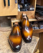 A pair of men's slip-on dress shoes with a glossy brown and amber gradient finish, displayed on a wooden workbench in a workshop setting.