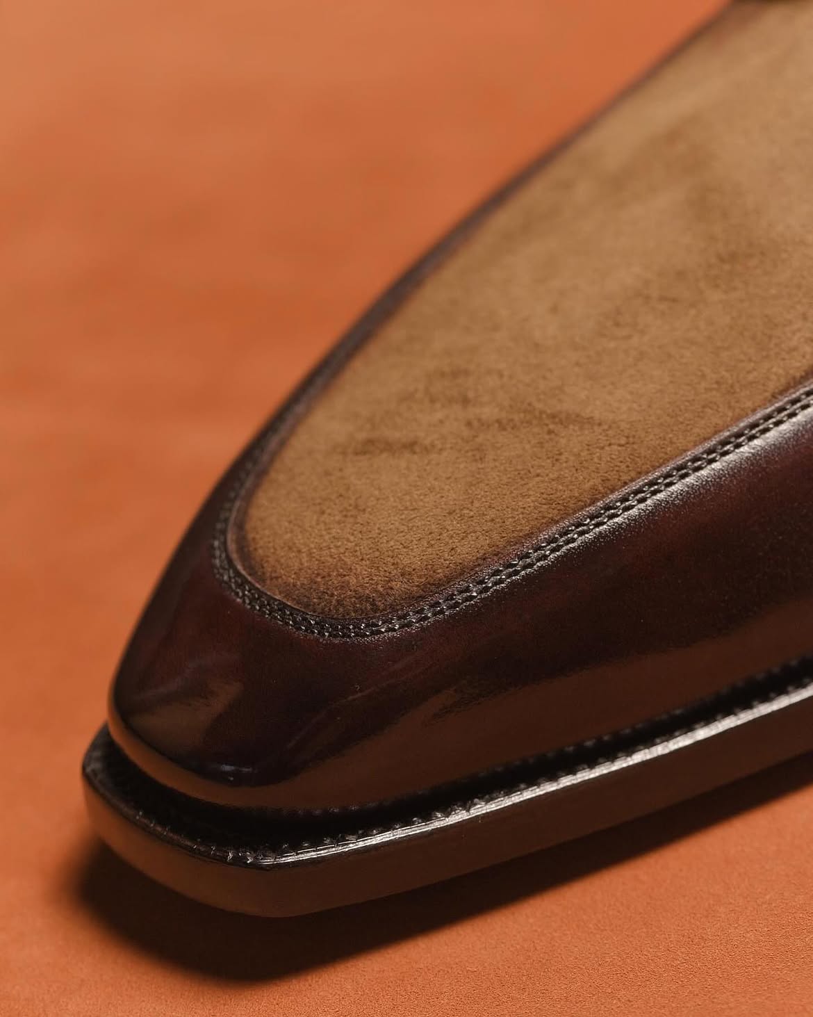 Close-up of a brown leather loafer toe showing a smooth dark brown vamp and a tan suede-like insole, set against a warm orange-brown background.