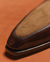Close-up of a brown leather loafer toe showing a smooth dark brown vamp and a tan suede-like insole, set against a warm orange-brown background.