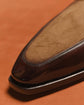 Close-up of a brown leather loafer toe showing a smooth dark brown vamp and a tan suede-like insole, set against a warm orange-brown background.