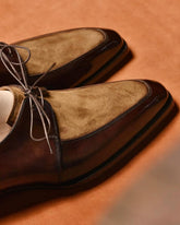 A pair of men's dress shoes with dark brown leather and tan suede panels, featuring thin gray laces, displayed on a warm brown textured surface.