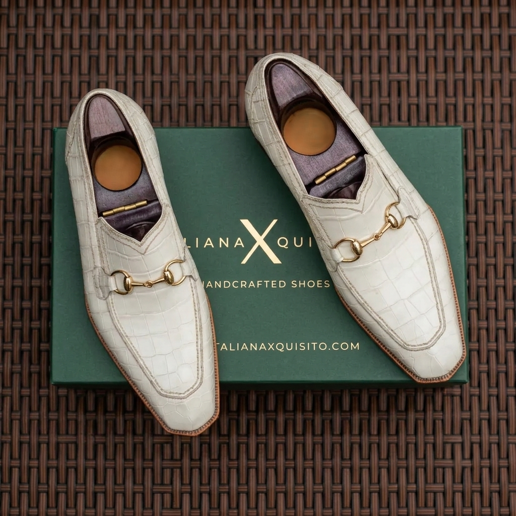 A pair of off-white crocodile-embossed loafers with gold horsebit detailing, displayed on top of a green shoebox on a brown woven mat background.