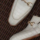 A close-up of a pair of off-white crocodile-embossed loafers with gold horsebit details, resting on a brown woven wicker surface.