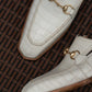 A close-up of a pair of off-white crocodile-embossed loafers with gold horsebit details, resting on a brown woven wicker surface.