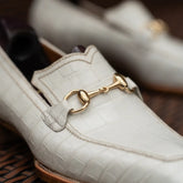A close-up of a white crocodile-embossed leather loafer with a gold horsebit detail, resting on a dark brown woven surface.