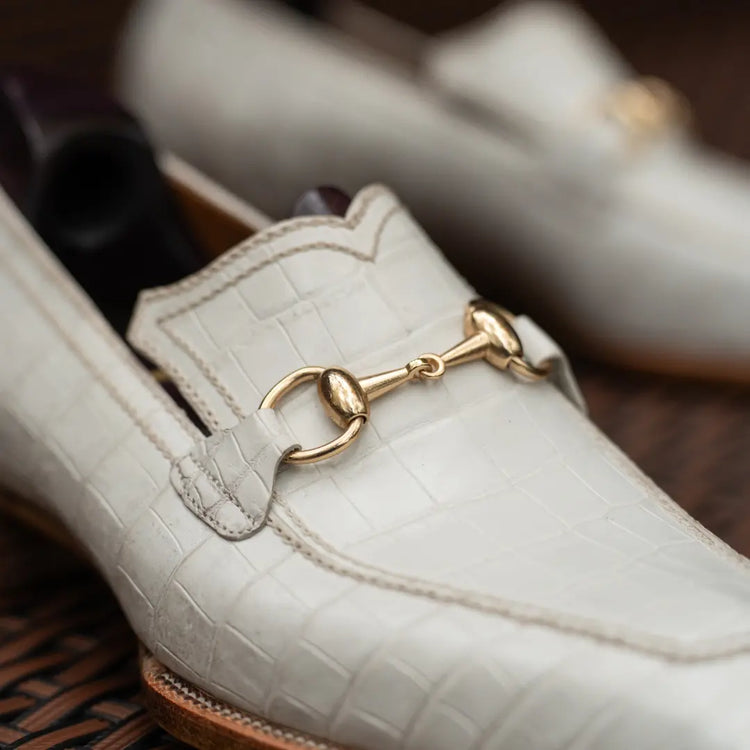 A close-up of a white crocodile-embossed leather loafer with a gold horsebit detail, resting on a dark brown woven surface.