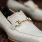 A close-up of a white crocodile-embossed leather loafer with a gold horsebit detail, resting on a dark brown woven surface.