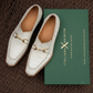A pair of white crocodile-embossed leather loafers with gold horsebit detailing, displayed next to a green 'ITALIANA QUISITO' shoebox on a dark brown woven mat background.